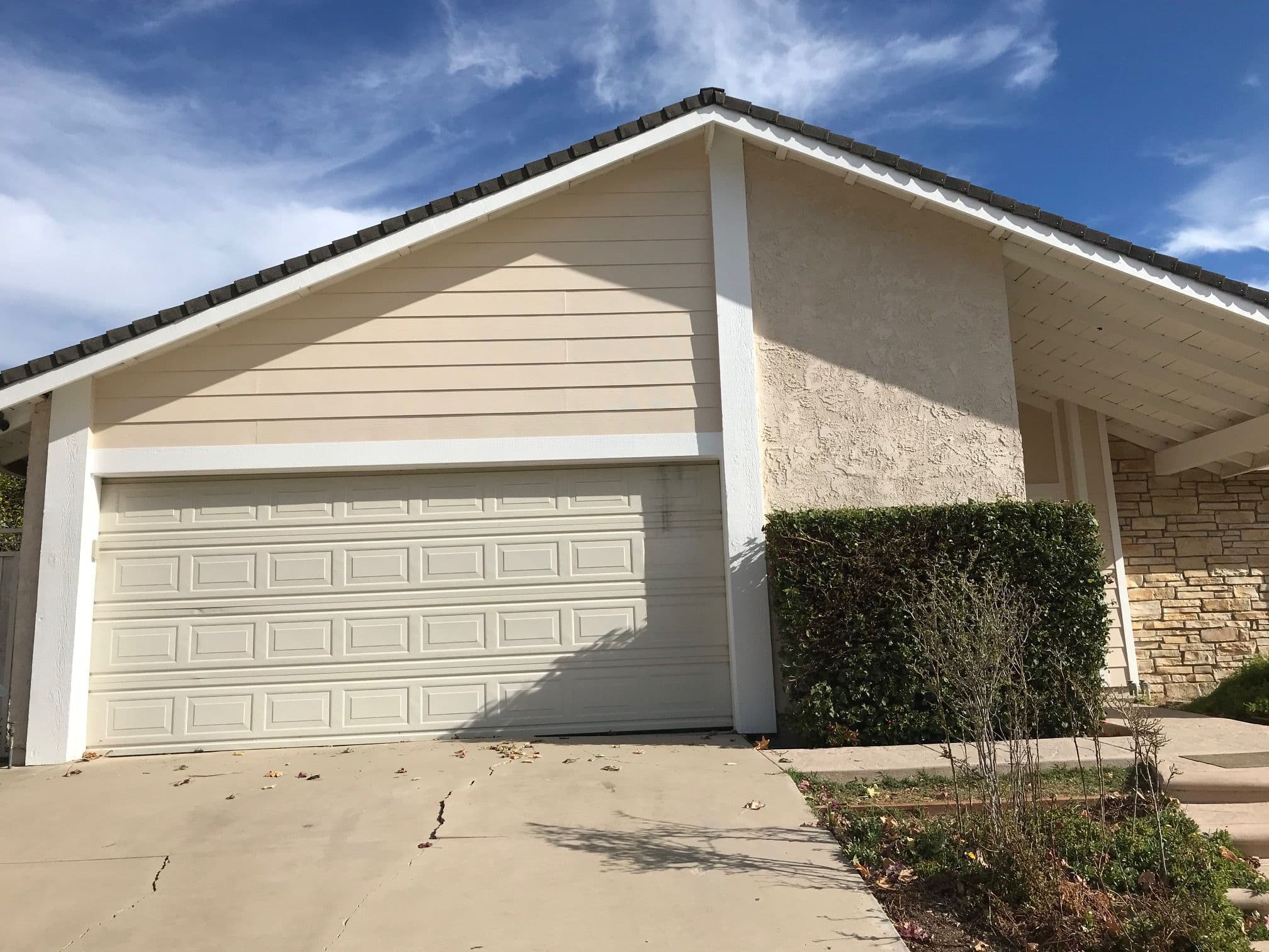 Single-story beige house with garage and manicured shrubs under a blue sky.