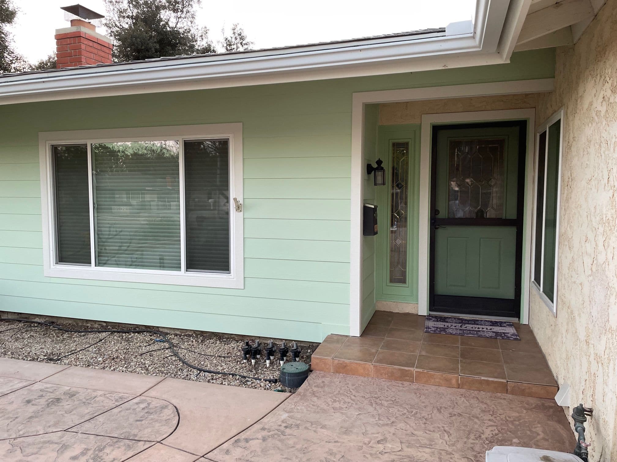 Home entrance with green siding, tile porch, and large windows surrounded by landscaping.