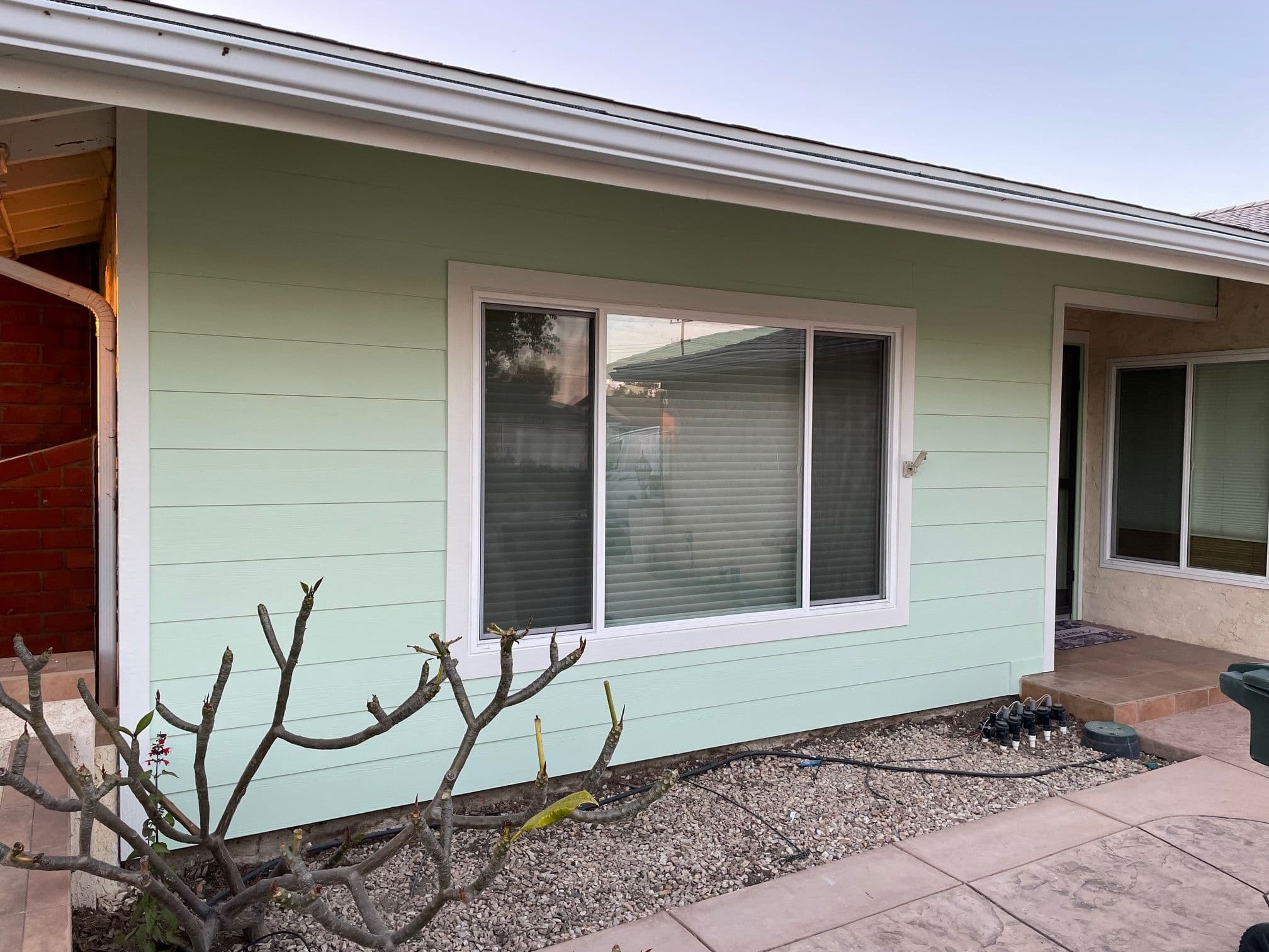 Light green house exterior with large window and modern siding, surrounded by landscaping.